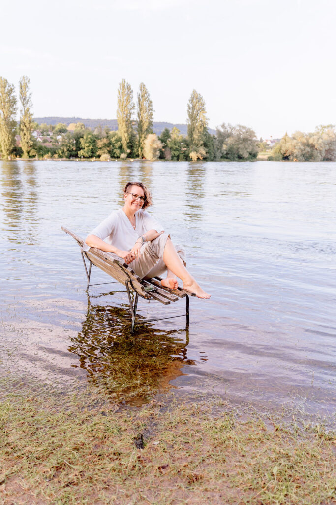 Corinna Coach für Nervensystemregulation und emotionale Balance sitzt auf einer Bank die in Wasser steht. Unabhängig von äusseren Umständen zufrieden.