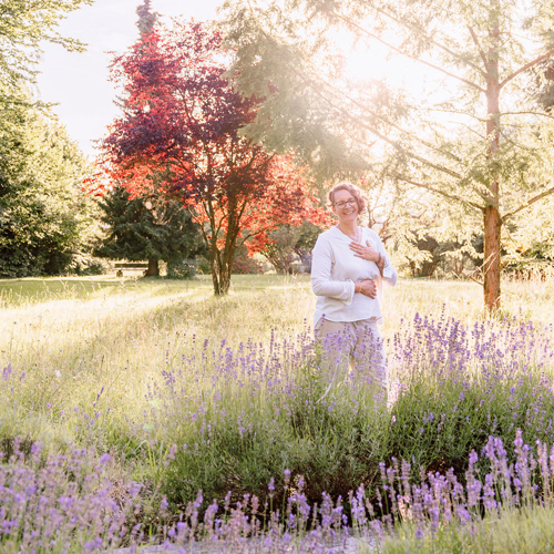 Corinna Fanni, Coach für Nervensystemregulation und emotionale Balance steht im Gras in der Abendsonne mit einem Lachen und den Händen auf dem Herz und Bauch.