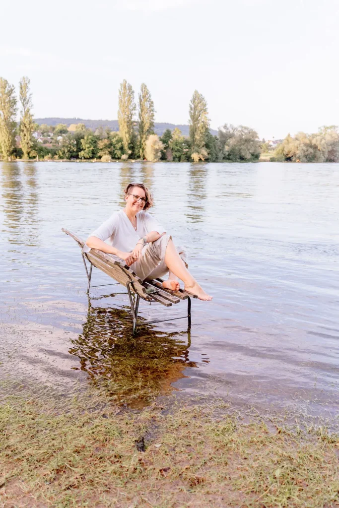 Corinna Coach sitzt auf einer Bank die in Wasser steht. Unabhängig von äusseren Umständen zufrieden.